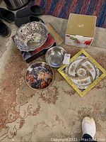 Top view photo showing the silverplate pierced bowl, clear glass bowl, decorative plates including round plate with multi-figure artistic scene, square plate with gold floral design, and small silver-tone bowl arranged on a carpeted floor.