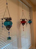 Three of the hanging glass candle holders visible, including two blue and one red, suspended from chains against window blinds.