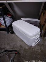 Full view of white Igloo cooler on the floor next to a shelf, showing the closed lid and overall shape.