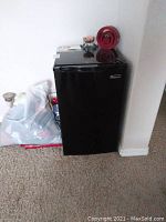 Full view of the black Magic Chef mini fridge against a wall on carpeted floor, showing size and external condition.