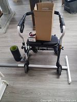 View of the Drive walker showing black padded seat and foam handles, two 5 pound red weights on the floor next to a black foam roller and a white plastic roller accessory, with a cardboard box on the seat.