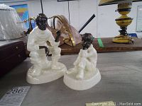 Two Minton porcelain and bronze figurines displayed on a table with other items in background. The larger male figurine is squatting, the smaller child figurine is seated.