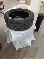 Stack of four black tires with visible tread, stacked one on top of another on a white plastic sack inside a room with wood floor.