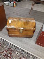 Front and side view of vintage wooden chest with ribbed yellow-orange metal lid, metal locking hardware, placed on floor with patterned rug.