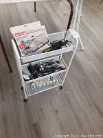 White metal rack on casters holding sewing machine box on top tier, black hair trimmers and cords on middle tier, and white hair rollers on bottom tier, on hardwood floor.