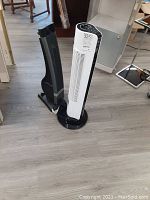 Two oscillating tower fans on a wood floor, one white with black sides, the other mostly black, shown side by side.
