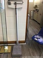 Full view of the vintage medical scale standing upright against white brick wall, showing the complete structure with platform and beam.