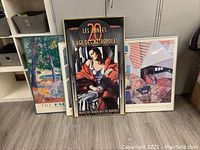 Three framed posters positioned standing on floor, showing varying frame sizes and styles, with mainly artistic prints including a colorful interior, an art deco woman, and impressionist landscape.