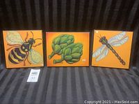 Three square plaques arranged side by side on a dark striped fabric background showing bee, artichokes, and dragonfly designs.