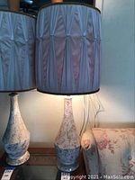 Lamp with neutral toned speckled base and light pink pleated fabric shade with gold trim on top edge, lamp illuminated.