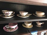 Six assorted vintage porcelain tea cup and saucer pairs on a black shelf, displaying various floral and gold detailed patterns.
