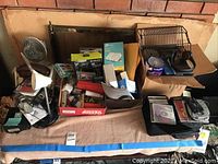 Full overview of the mixed electronics lot showing the fan, lamp, telephones, CD cases, and various packed items on a table covered with a beige towel in front of a brick wall.