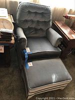 Front angle of the blue velvet upholstered chair with matching ottoman in front. Chair has a tufted cushion back and padded armrests; ottoman cushioned and matching.