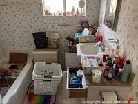 View of bathroom sink area with various personal care bottles, candles, tissues, baskets and trash cans around and under the sink.