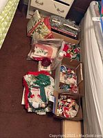 Photo showing boxes and bags of assorted Christmas fabric decorations and ornaments on a carpeted floor near a bed.