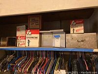 Photo of various film developing tanks and boxes on a shelf above hanging clothes, includes FR Corporation boxes and a Bakelite box.