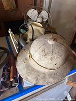 View of white pith hat on table near antique fan showing exterior of hat and fan behind it