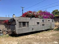 Side view of 1953 Mobile Trailer Company trailer exterior showing rectangular metal body, older worn tires on blocks, shallow windows, laundry pole, and surrounding yard.
