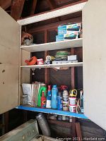 Cabinet with three shelves showing various plumbing, hardware, and cleaning supplies including boxes of digital TV tuners on the top shelf, coiled tubing, and spray bottles on middle and bottom shelves.