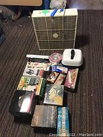 Photo shows a large beige box fan and an assortment of kitchen tools including boxed items, a white container with a black handle, and other kitchen gadgets arranged on a brown carpeted floor.