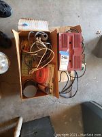 Box with vintage red automotive battery charger with wires, Sears utility pump and Pittsburgh transfer pump in packaging.
