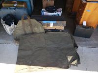 Photo showing green military canvas tarp spread out partially with a folded duffel bag nearby. The tarp has metal grommets on corners. The duffel bag is large, olive green with handles.