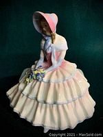 Front view of Royal Doulton porcelain figurine seated young woman in pink ruffled dress and bonnet holding flowers.