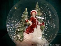 Interior view showing porcelain lady in red coat holding gifts among fir trees with faux snow inside glass globe