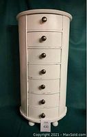 Full front view of tall narrow white side table with seven drawers with black round knobs on green background.
