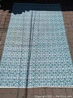 Full view of rectangular turquoise and white area rug laid flat on outdoor brick surface, showing entire rug shape, pattern, and size impression.