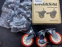 Photo shows the majority of 8 caster wheels, including 3 with red polyurethane wheels and several packed in plastic bags next to a cardboard box.