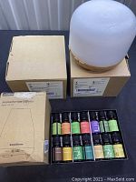 Display of one diffuser with white dome and wooden base alongside diffuser boxes and 12 essential oil bottles in black tray.