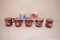 Seven vintage mugs shown in lineup, five maroon with white Native American scenes, two white with red/blue Davy Crockett scenes.