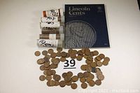 Wide shot showing five labeled coin tubes, a stack of pennies spread on table, and an empty Lincoln Cent coin album with a large penny image on the cover.
