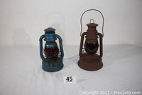 Photo showing both lanterns side by side, the blue painted Dietz Little Wizard with ruby red globe on left, rusty brown Dietz Little Giant with red globe on right.