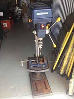 Front angle view of the Mastercraft drill press showing the head, control lever with yellow grips, and the base stand with visible signs of rust and wear.