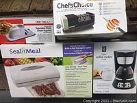 Photo showing the boxed items including the One Touch automatic vegetable slicer, Chef'sChoice blade sharpener, Seal a Meal vacuum sealer, and black digital coffee maker.