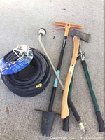 Photo showing entire set of garden tools displayed on concrete, including the rolled black rubber hose, Fiskars branch trimmer, axe with wooden handle, green handled pruner, and garden hole planter with metal pole.