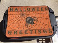Closed orange tote bin with Halloween Greetings doormat on top showing spider web and spider design.