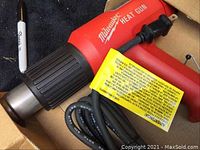 Photo of the Milwaukee heat gun in the box showing the red gun with black nozzle and cord with warning label attached.