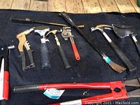 Photo showing an array of hand tools laid out on a black carpet including bolt cutters, rivet gun, hammers, pipe wrench, pry bar, and axes.