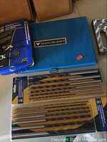 Mastercraft blue plastic case with tap and die set, assorted drill bits in packaging, staple gun, and hammer on floor.