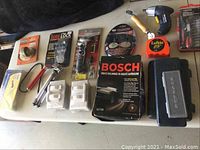 Wide view of the lot showing all items on a table including Bosch ignition cables kit, Lufkin tape measure, mini drill, Sure Lock device, sharpening attachment and mini tripod.
