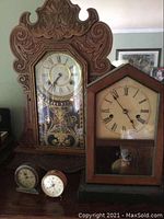 Wide shot showing all four clocks: the large Waterbury mantle clock with ornately carved wood and decorative glass, second wooden mantle clock with simpler design, and two smaller tabletop clocks.