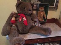 Large Gund brown plush bear seated on table with smaller Dickey bear with red shorts on its arm and a light brown Dickey bear in display case behind.