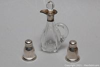 Full view of two sterling silver salt and pepper shakers flanking the glass cruet with silver neck and stopper, displayed against a white background.