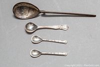 Four sterling silver spoons laid out on flat surface; one long spoon at top, three small decorative salt cellar spoons below.