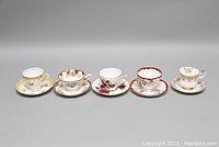 Five vintage teacups and matching saucers in a row, showing floral patterns and gold trim.