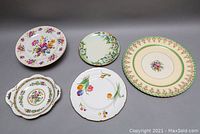 Top view of five assorted plates and one double handle bowl arranged on gray surface. Various floral patterns and ornate designs visible.