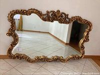 Front view of large rectangular mirror with ornate gold-colored carved frame on tiled floor